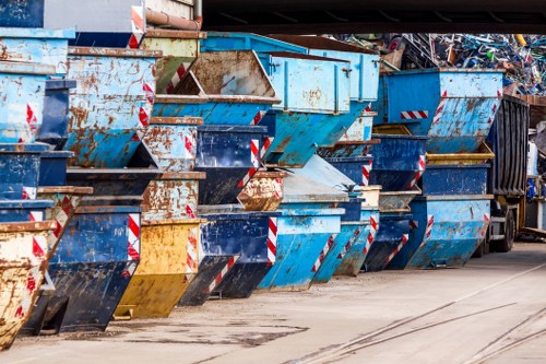 A row of large metal commercial waste bins, predominantly painted in shades of blue with some showing rust and wear, lined up on a paved area, likely part of a waste disposal or recycling facility. The bins are positioned side by side, with their open tops and front panels visible, some displaying chipped paint and signs of usage. Behind the bins, there are stacked piles of discarded electronic waste and other debris, indicating a busy environment related to rubbish removal or clearance services. The ground beneath the bins is concrete, with visible tire marks and some liquid stains, suggesting frequent vehicle access. The overall scene reflects an organised collection point for bulk rubbish, typical of waste management operations found in an urban or suburban setting, supporting companies like Barnes House Clearance in their domestic or commercial rubbish clearance activities in Barnes or nearby areas.