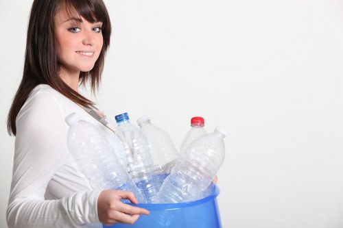 A young woman with shoulder-length brown hair and a white long-sleeved top is holding a blue plastic bucket filled with several empty plastic bottles of various sizes, mostly transparent with some having red or blue caps. The bottles are made of clear PET material and are loosely arranged within the bucket, with some leaning against the sides. The woman stands against a plain white background, smiling gently and looking slightly to the camera's left. The image captures a clean, indoor environment and emphasizes the object’s focus on waste collection and recycling. The scene relates to rubbish disposal, aligning with services offered by Barnes House Clearance for proper waste management and recyclable rubbish removal, subtly supporting local SEO for London-area rubbish clearance services.