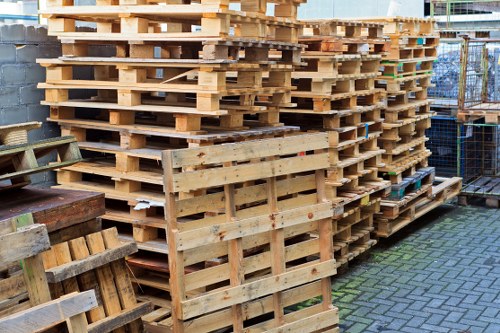 A large stack of untreated wooden pallets arranged neatly outdoors on a paved surface, with some pallets leaning against a concrete wall and others stacked in multiple columns. The pallets are light-colored, showing natural wood grain and rough textures typical of shipping or storage pallets. In the background, a chain-link fence and a partially visible building suggest an industrial or storage yard environment, likely at a waste or rubbish removal site. The scene is evenly lit, with no noticeable shadows, and the pallets are in good condition, with some showing minor signs of wear. This setup is indicative of waste preparation for disposal or recycling, aligning with services offered by Barnes House Clearance in the context of rubbish removal and clearance in the local area.